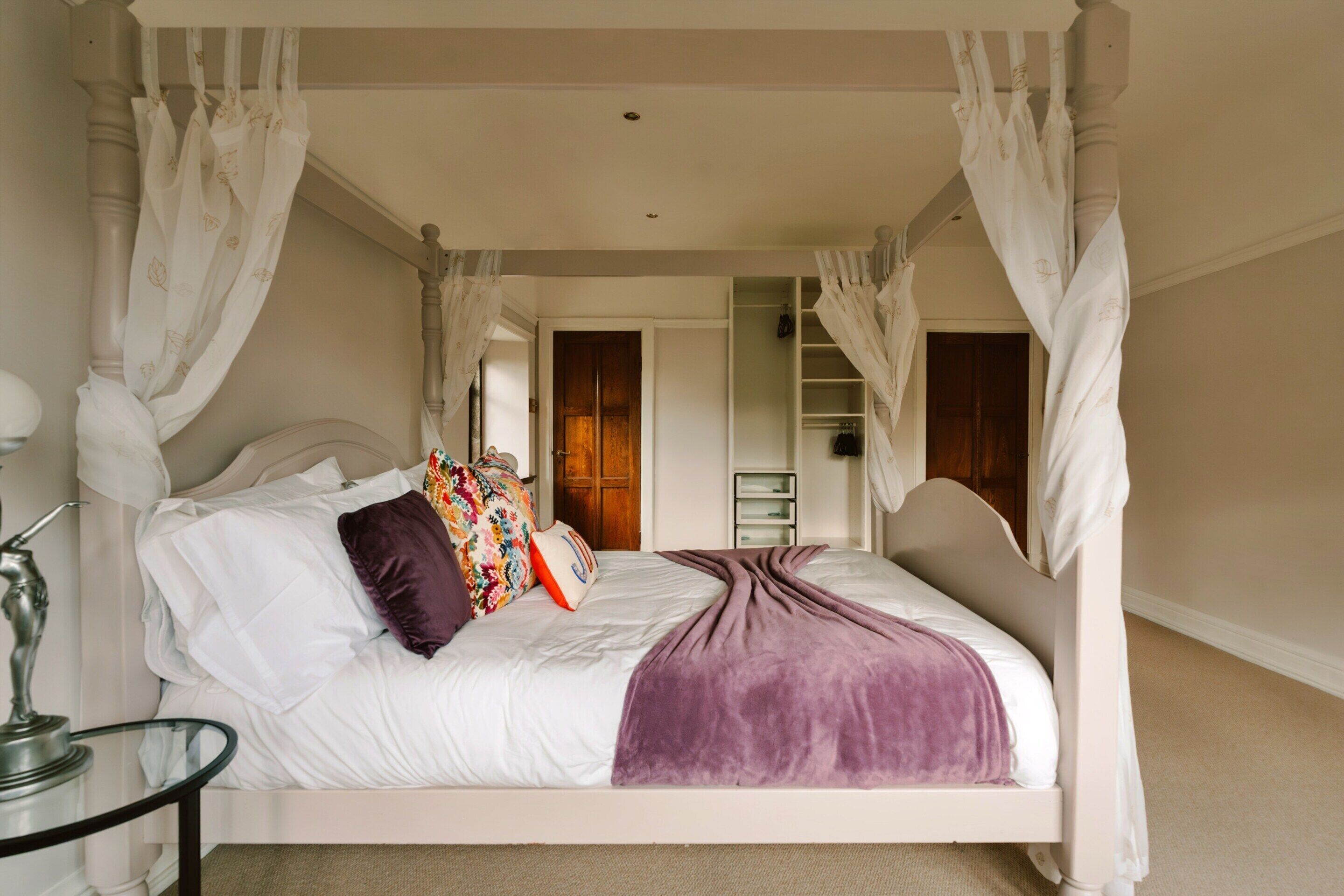 A four-poster canopy bed with a white headboard, multiple pillows, and a purple throw blanket.