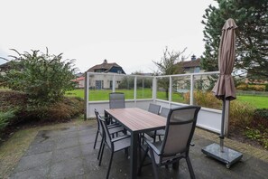 Ein überdachter Patio-Bereich mit Tisch und Stühlen, einem Metallschirm und Blick auf ein Haus und einen Garten.