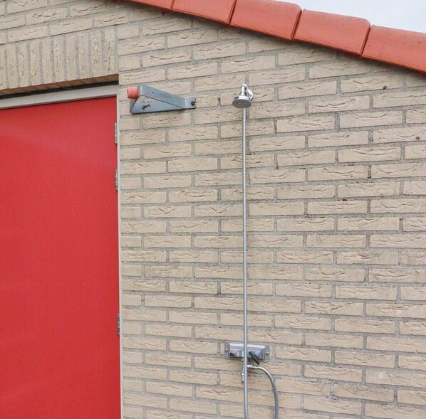 Eine Ziegelwand mit einem vertikal verlaufenden Metallrohr, eine rote Tür und eine kleine Metallkasten mit angeschlossenem Schlauch.