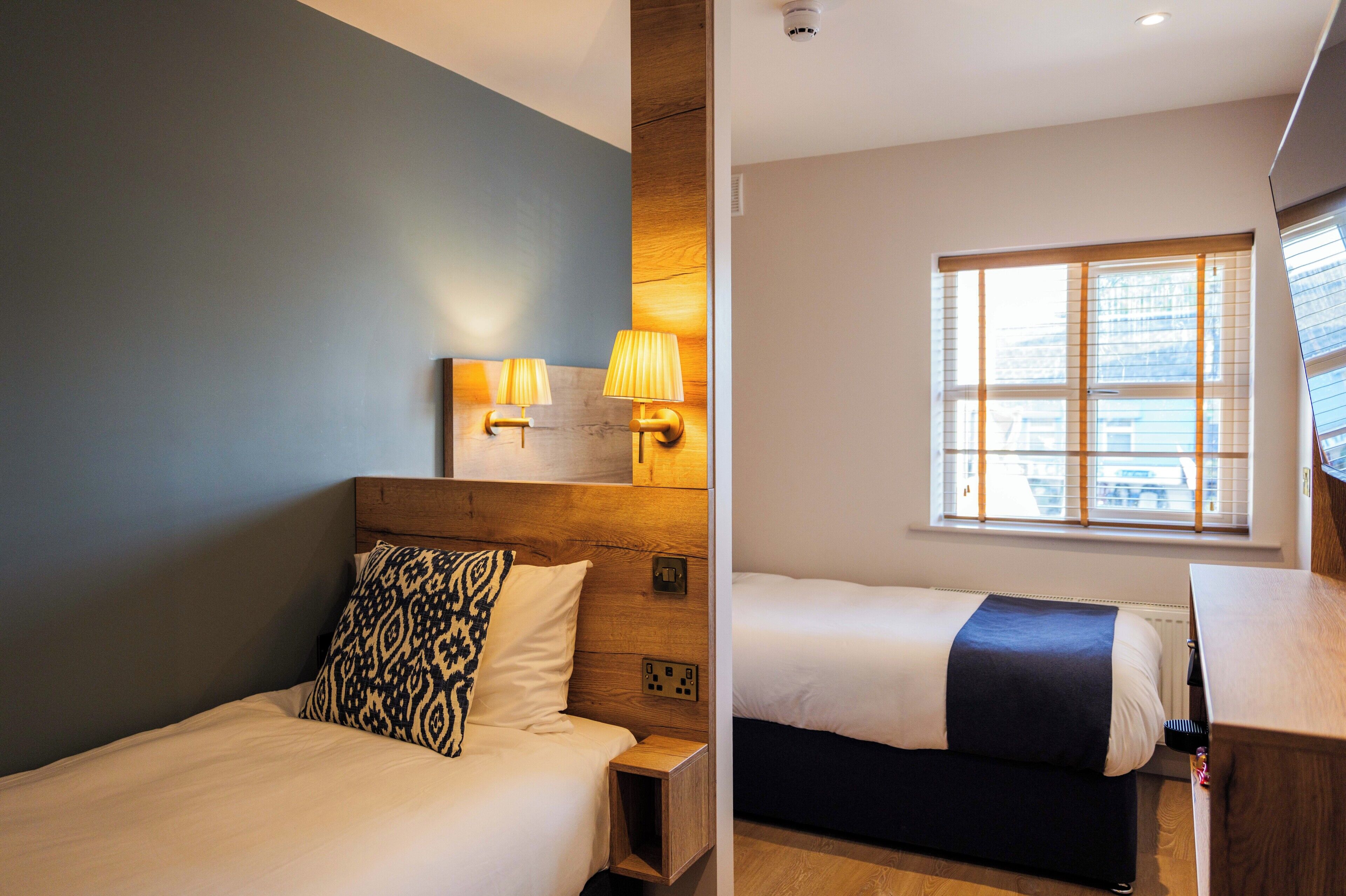 A hotel room with a bed, bedside table, lamp, and a window with blinds.