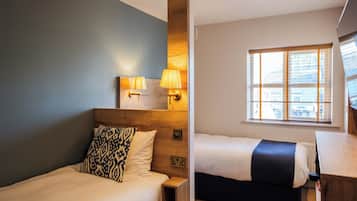 A hotel room with a bed, bedside table, lamp, and a window with blinds.