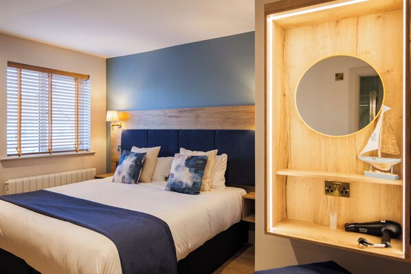 A hotel room with a bed, a nightstand, a lamp, a mirror, and a small shelf with a hairdryer.