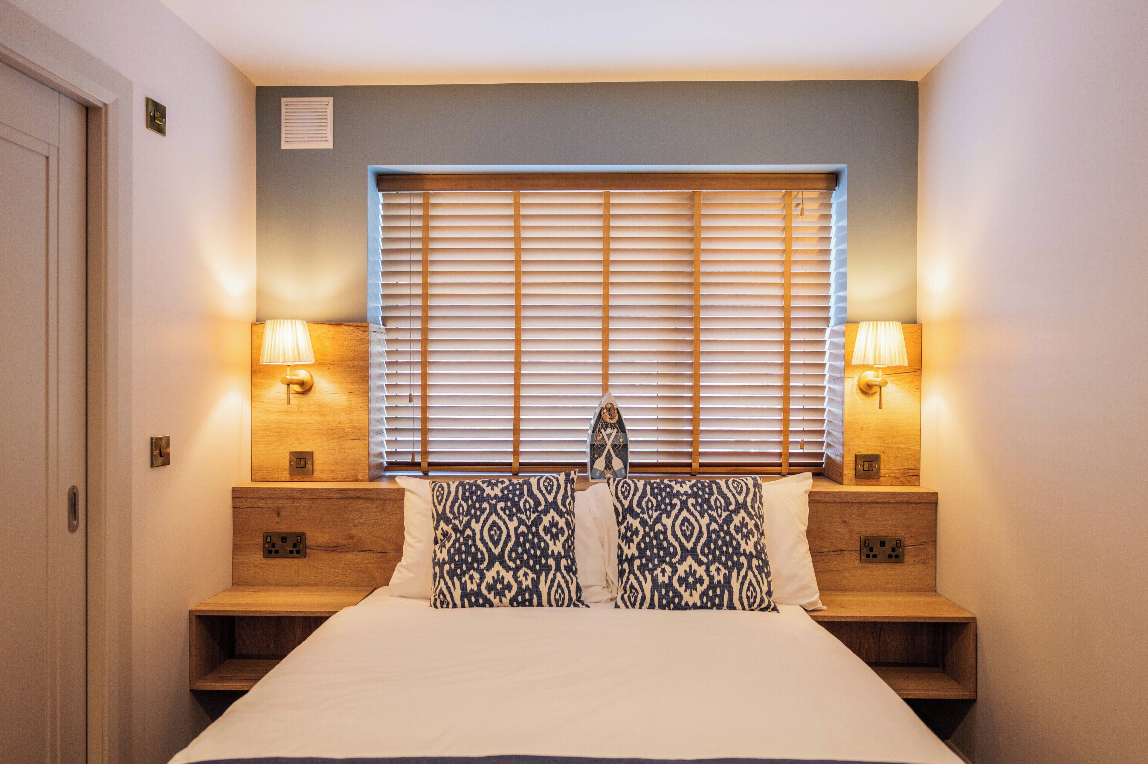 Un dormitorio con cama, dos mesitas de noche, dos lámparas, una ventana con persianas y un interruptor de luz en la pared.
