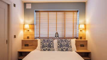 Un dormitorio con cama, dos mesitas de noche, dos lámparas, una ventana con persianas y un interruptor de luz en la pared.