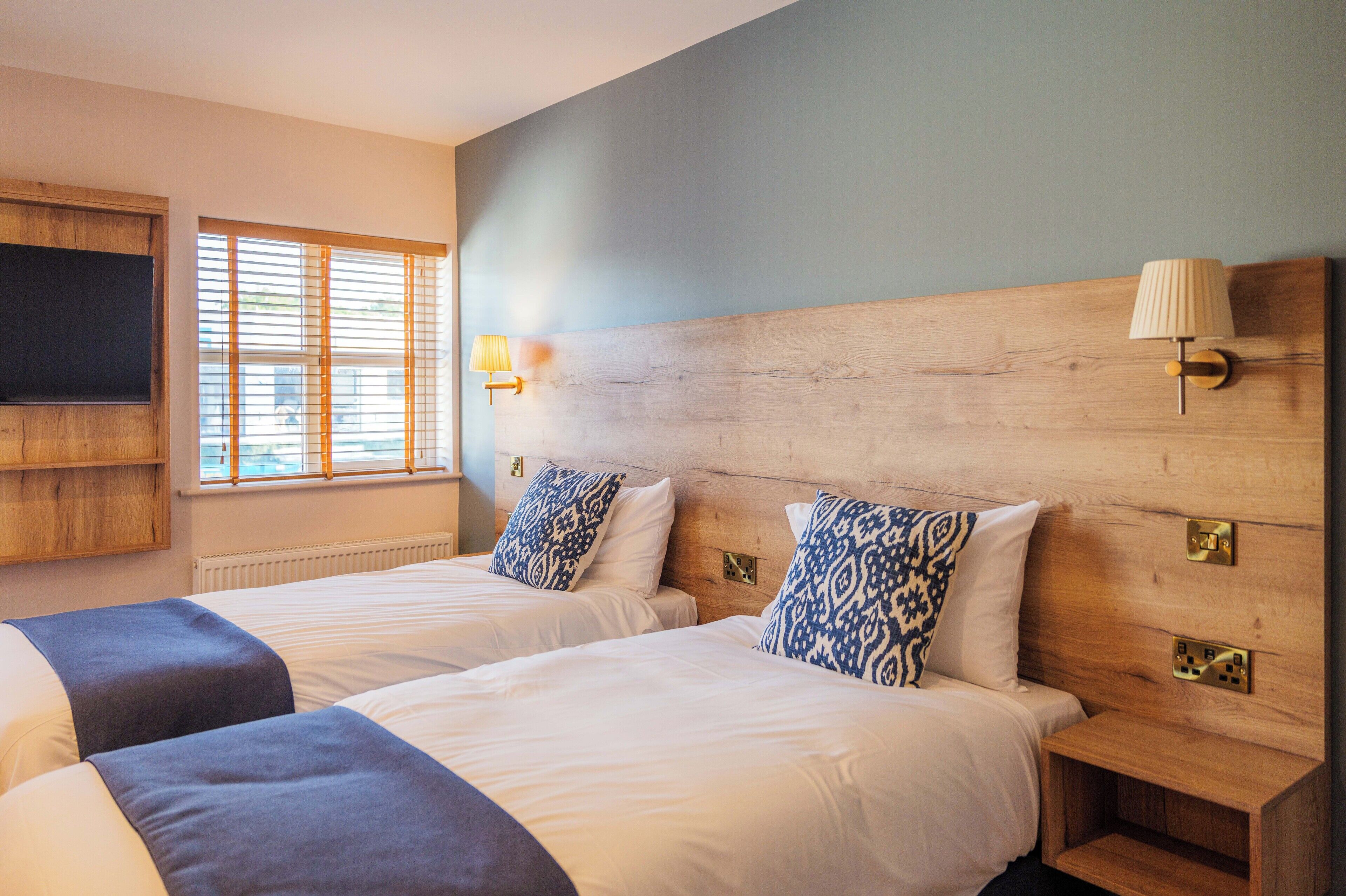 A hotel room with two beds, a television mounted on the wall, and a window with blinds.