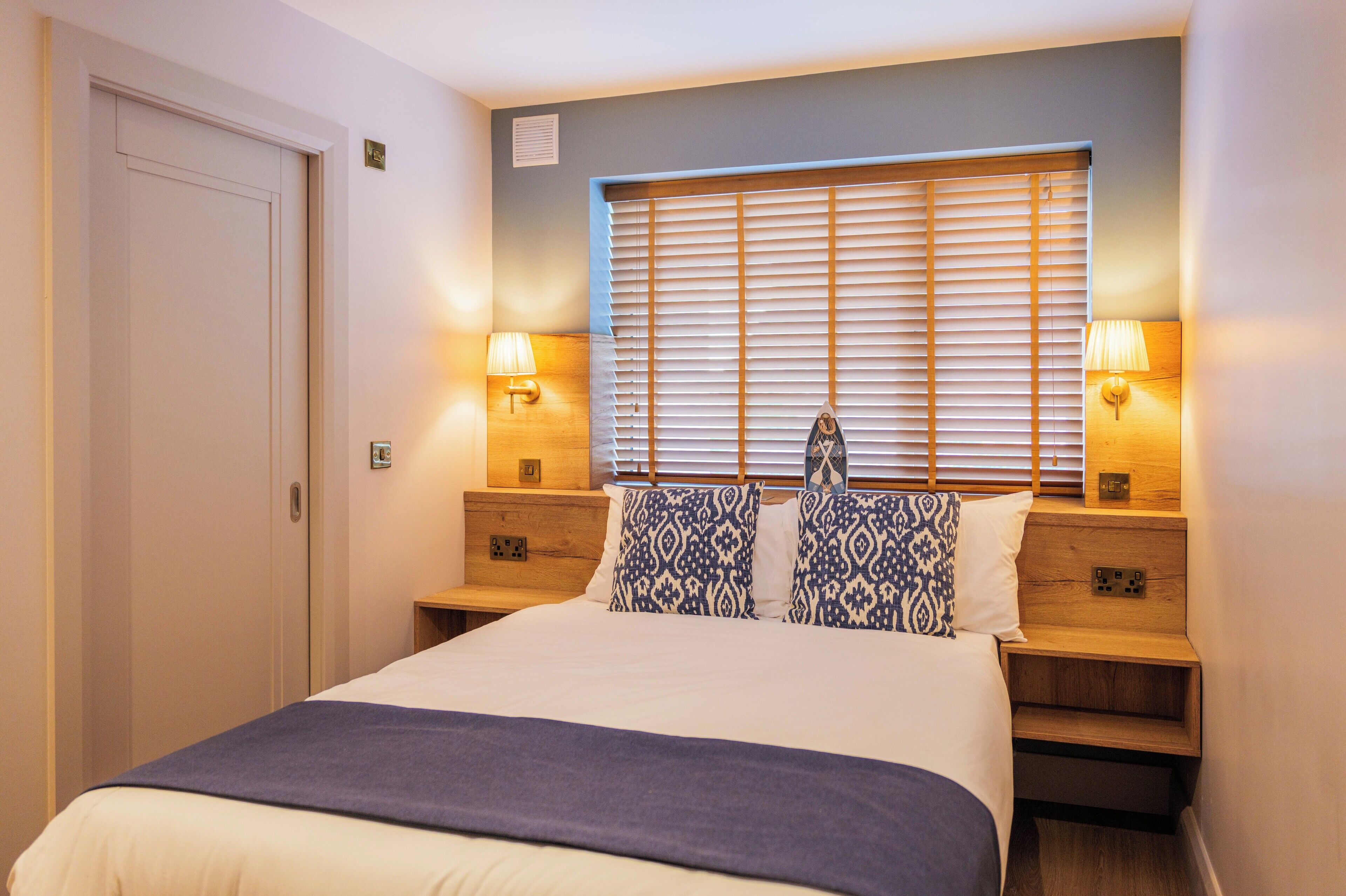 A hotel room with a bed, two bedside tables with lamps, a window with blinds, and a door.