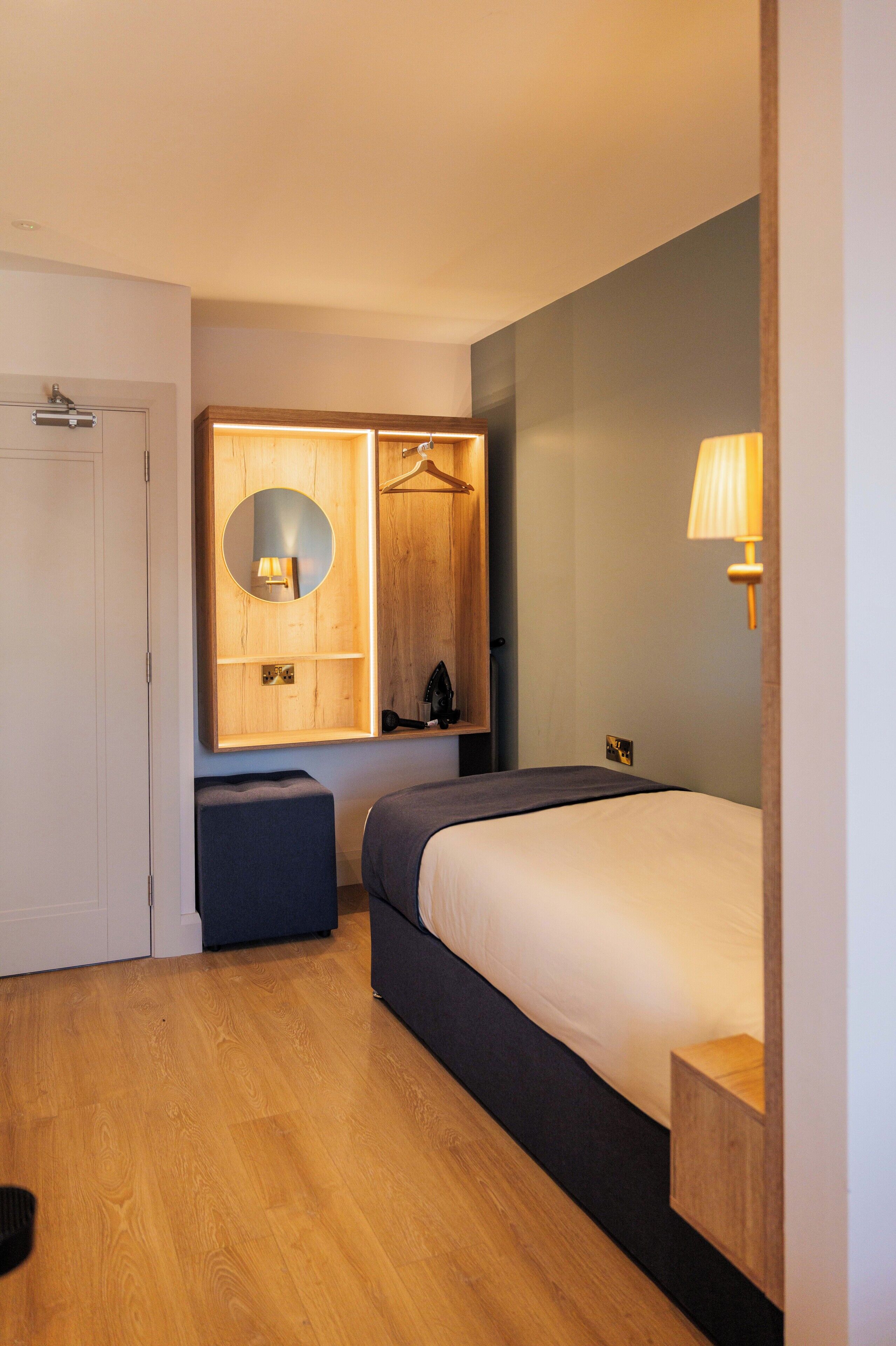 Una habitación de hotel con cama, mesita de noche, lámpara, espejo y armario.