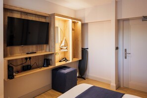 A hotel room with a large flat-screen TV mounted on a wooden wall, a small sailboat decoration, a black office chair, and a door with a handle.