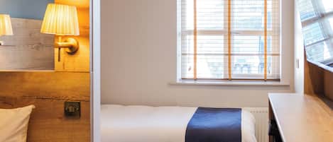 A hotel room with a bed, bedside tables, a lamp, a window with blinds, and a wooden floor.