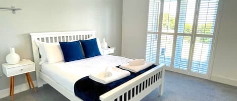 Ein Schlafzimmer mit einem weißen Bett, blauen Kissen, einem weißen Nachttisch und einem großen Fenster mit Jalousien.