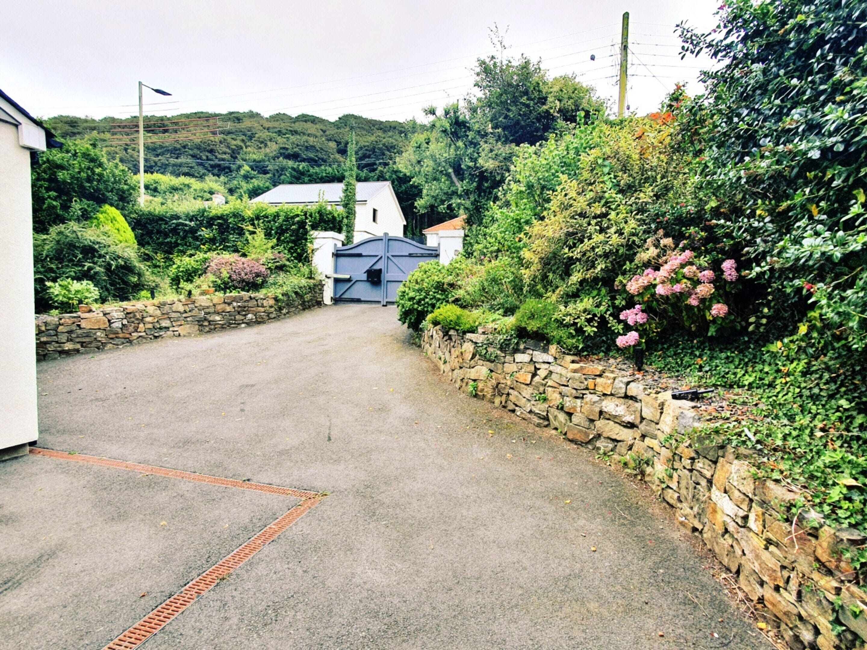 A paved driveway leading to a gated entrance with a stone wall on one side and a building with a sloped roof in the background.