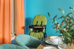 Ein Zimmer mit einem grünen Polsterstuhl, einem Kinderwagen, einem Fenster mit orangefarbenen Vorhängen und einem Tisch mit Tellern und einem Glas.