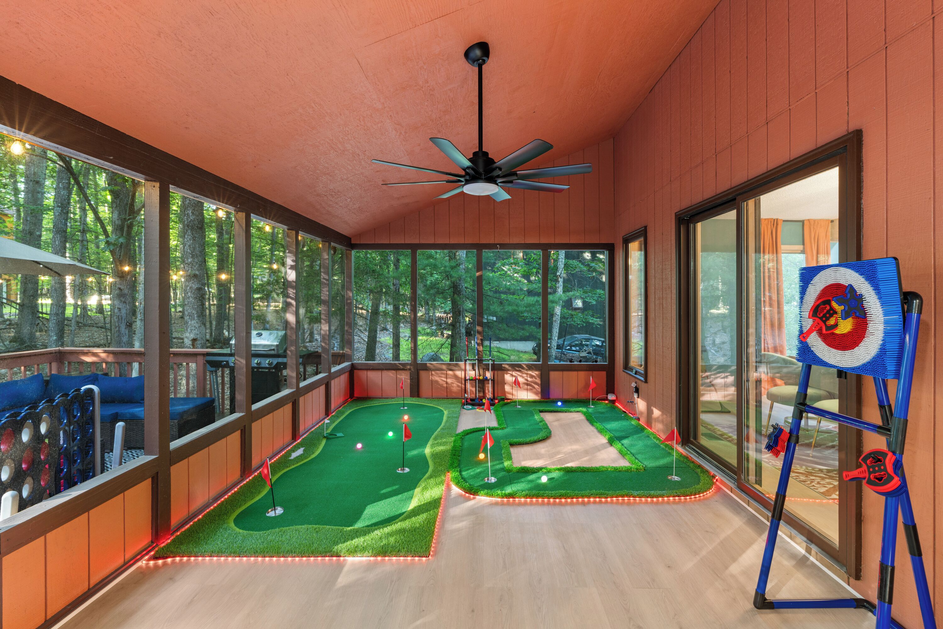 A mini-golf setup with a ceiling fan, large windows, and a view of the surrounding forest.
