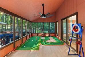 Ein Minigolfplatz mit Deckenventilator, großen Fenstern und Blick auf den umliegenden Wald.