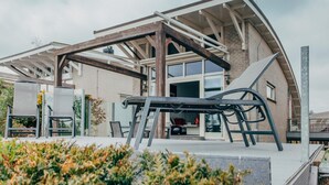 Een modern gebouw met een houten constructie, grote ramen en een overdekt buitengebied met zitplaatsen.