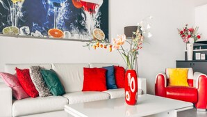 Een moderne woonkamer met een witte bank, kleurrijke kussens, een rode vaas met bloemen en een groot abstract schilderij erboven.