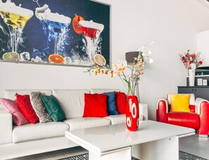 A modern living room with a white sofa, colorful cushions, a red vase with flowers, and a large abstract painting above the sofa. - Deluxe Dike Villa With Sauna for 6 (Makkum)