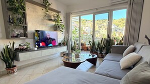 A modern living room with a large sofa, a glass coffee table, and a flat-screen TV. There are multiple potted plants and a view of a hillside through large windows. - Spacious SeaView Apartment in the heart of Saranda (Saranda)