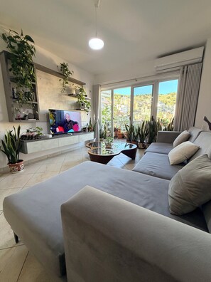 A modern living room with a large sofa, a glass coffee table, and a flat-screen TV. There are multiple potted plants and a view of a hillside through large windows. - Spacious SeaView Apartment in the heart of Saranda (Saranda)