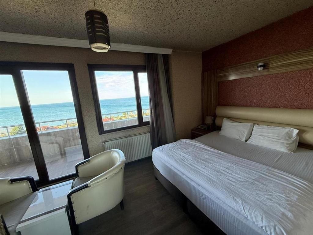 A hotel room with a bed, a chair, a nightstand, and a view of the sea through large windows.