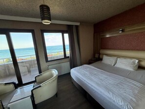 A hotel room with a bed, a chair, a nightstand, and a view of the sea through large windows.