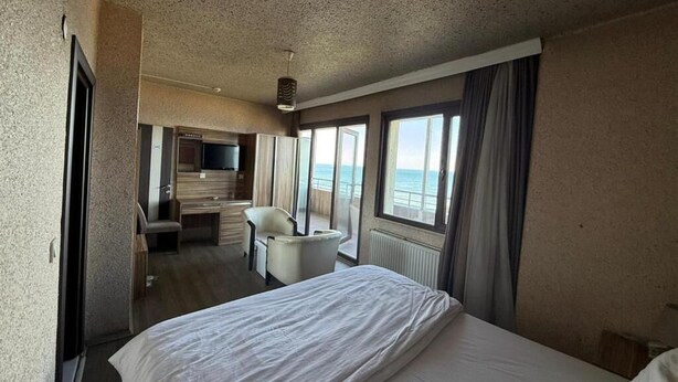 A hotel room with a bed, a desk, a chair, a TV, and large windows with curtains. - otel ustabaş (Hopa)