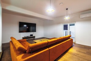 A modern living room with a large orange sofa, a flat-screen TV, a wooden coffee table, and pendant lighting.