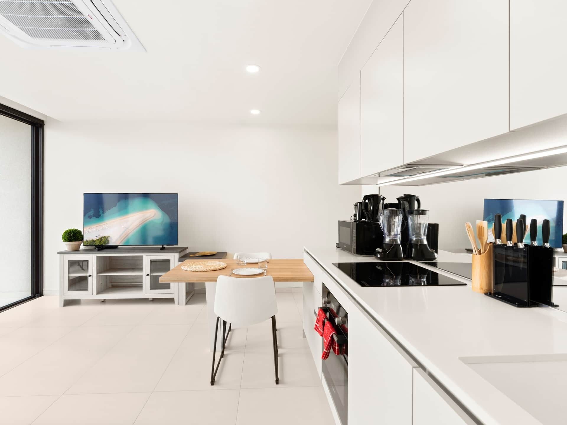 A modern kitchen with white cabinetry, a dining area with a wooden table, and a large flat-screen TV.