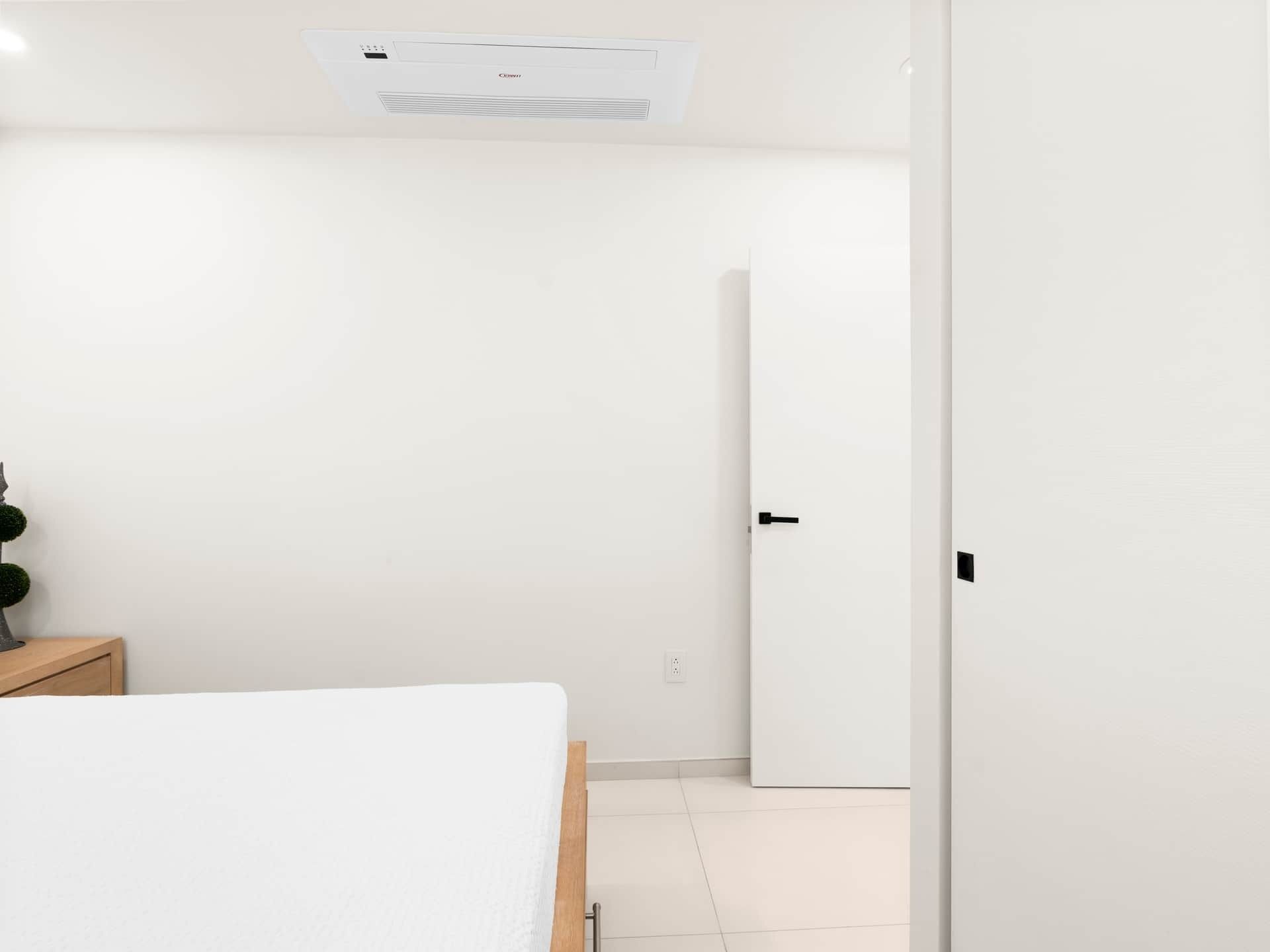 Une chambre d’hôtel minimaliste comprenant un lit, une table de chevet, un climatiseur fixé au mur et une porte.