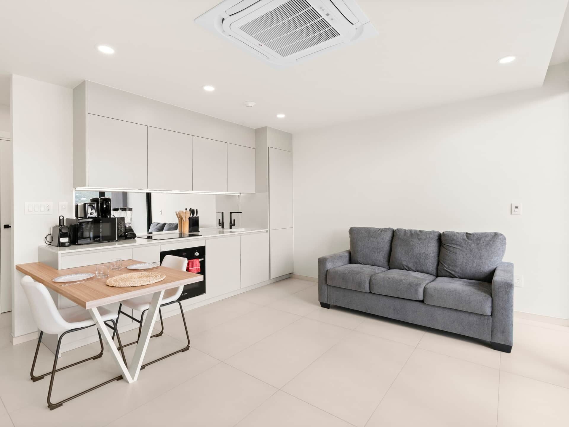 A modern kitchen with white cabinetry, a dining table with chairs, and a grey sofa in a bright, minimally furnished living area.