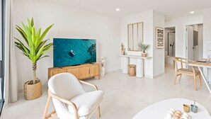 Uma sala de estar moderna com uma grande televisão de tela plana, uma poltrona confortável, uma área de jantar e uma cozinha visível ao fundo.