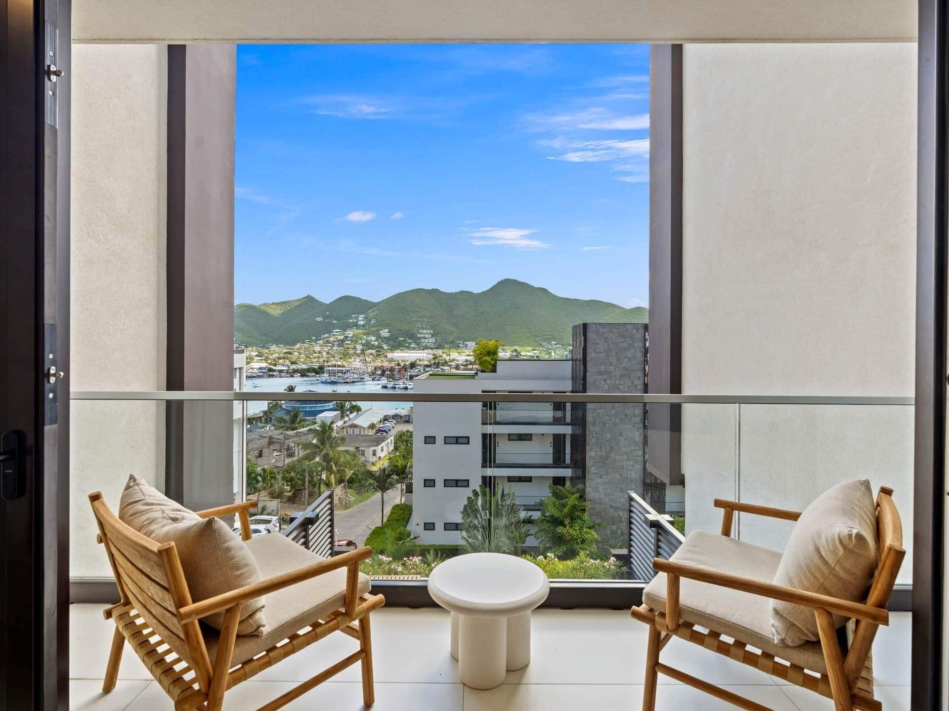 A balcony with two wicker chairs and a small table, overlooking a cityscape with mountains in the distance.