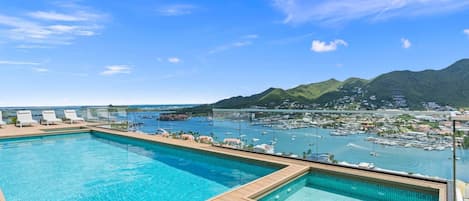 A swimming pool with clear blue water, surrounded by wooden deck and lounge chairs, overlooking a marina with boats and a coastal town.