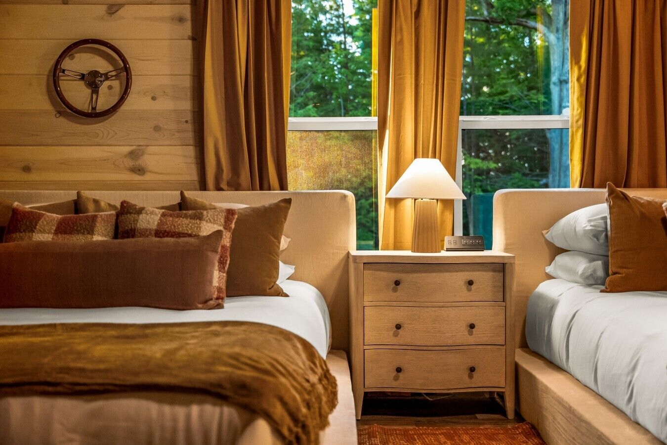Une chambre d’hôtel avec deux lits, une tête de lit en bois, une table de chevet avec une lampe et une fenêtre avec des rideaux.