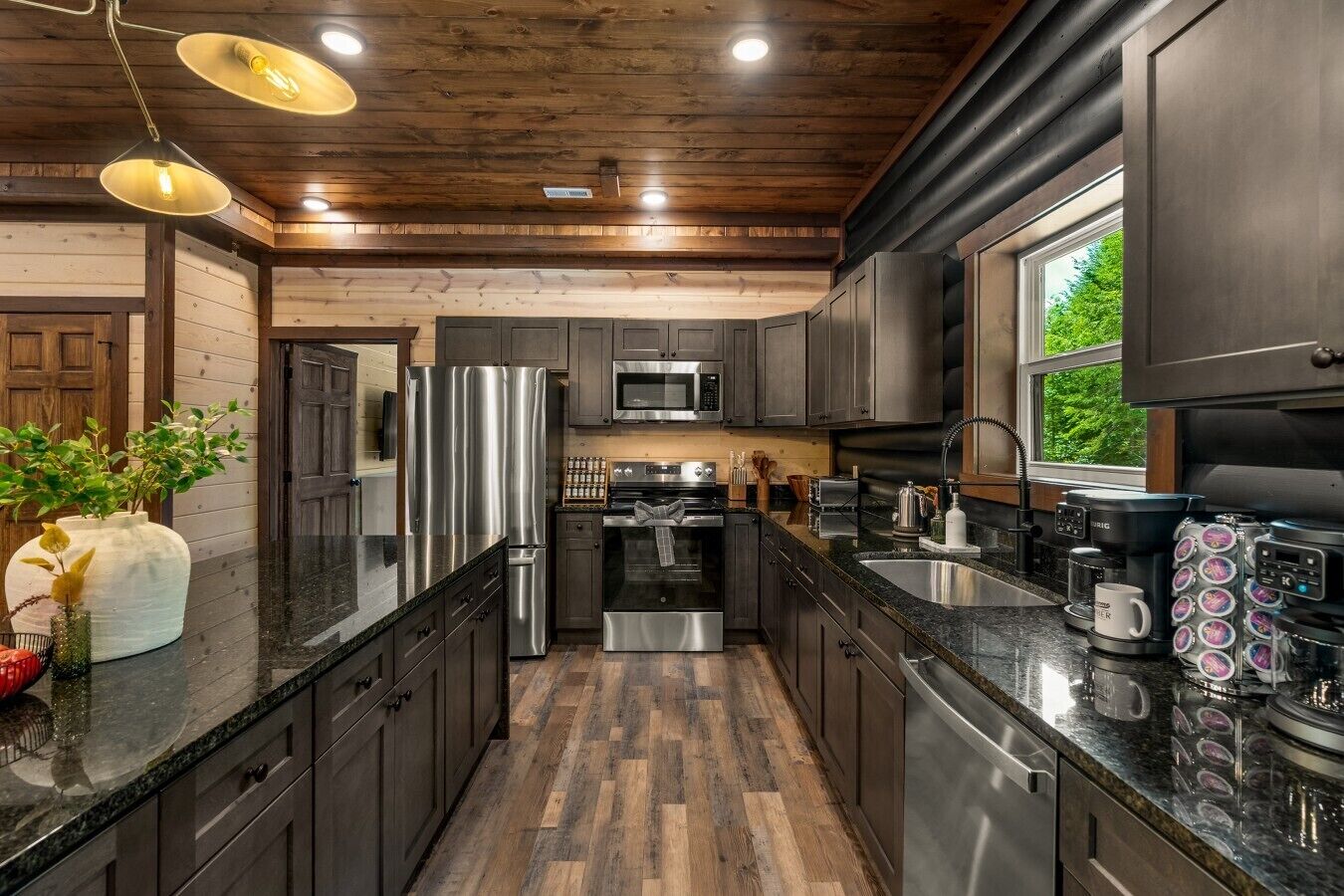 A modern kitchen with stainless steel appliances, wooden flooring, and a large window providing natural light.