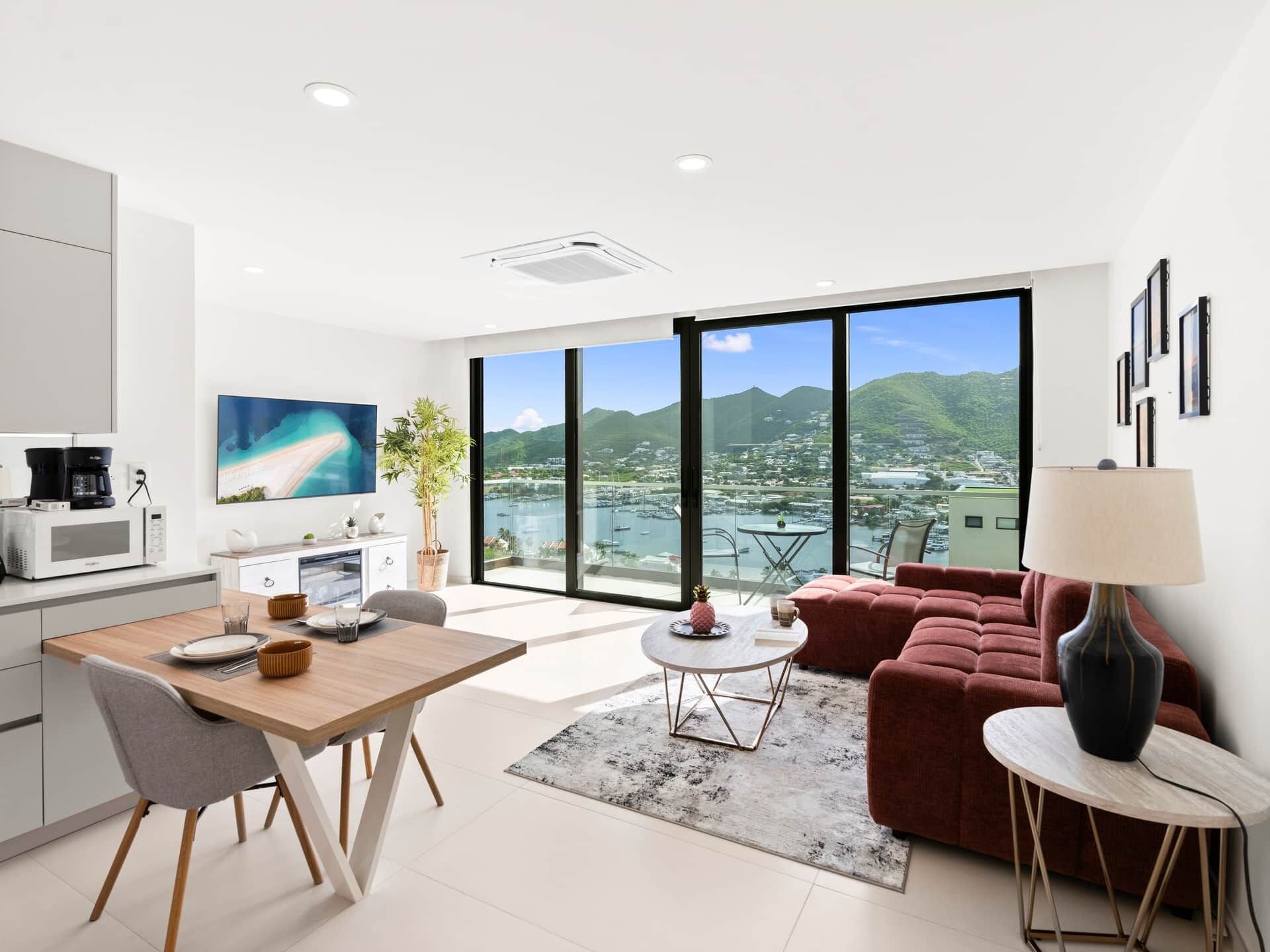 Un espacio habitable moderno con cocina, comedor y un amplio ventanal con vistas a un puerto deportivo y montañas.