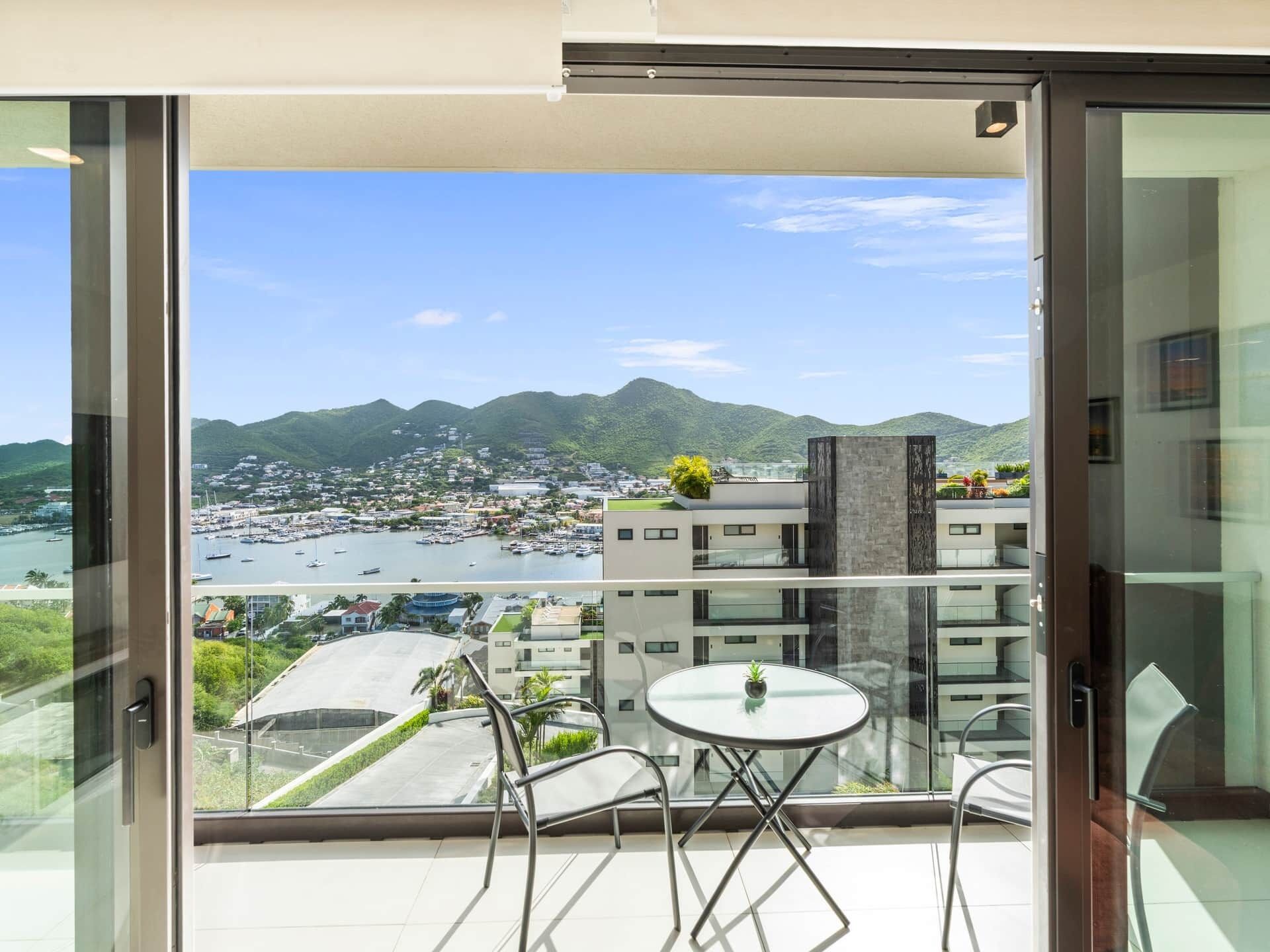 En balkon med glasdør, et bord og stole, der kigger ud over en lystbådehavn og et bjergrigt landskab.