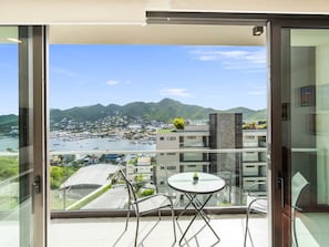 En balkon med glasdør, et bord og stole, der kigger ud over en lystbådehavn og et bjergrigt landskab.