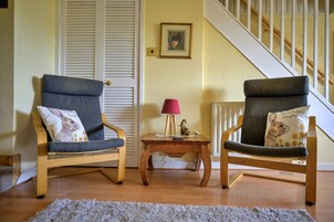 Two wooden chairs with grey cushions, a small wooden table with a lamp, and a staircase in the background.