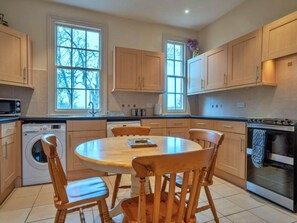 A kitchen with wooden cabinets, a washing machine, a dining table with chairs, and a stove. - Charming Derbyshire Cottage (Ashbourne)