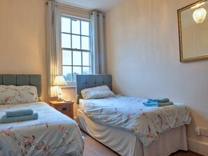 A bedroom with two beds, a window with curtains, a bedside lamp, and a mirror on the wall. - Charming Derbyshire Cottage (Ashbourne)