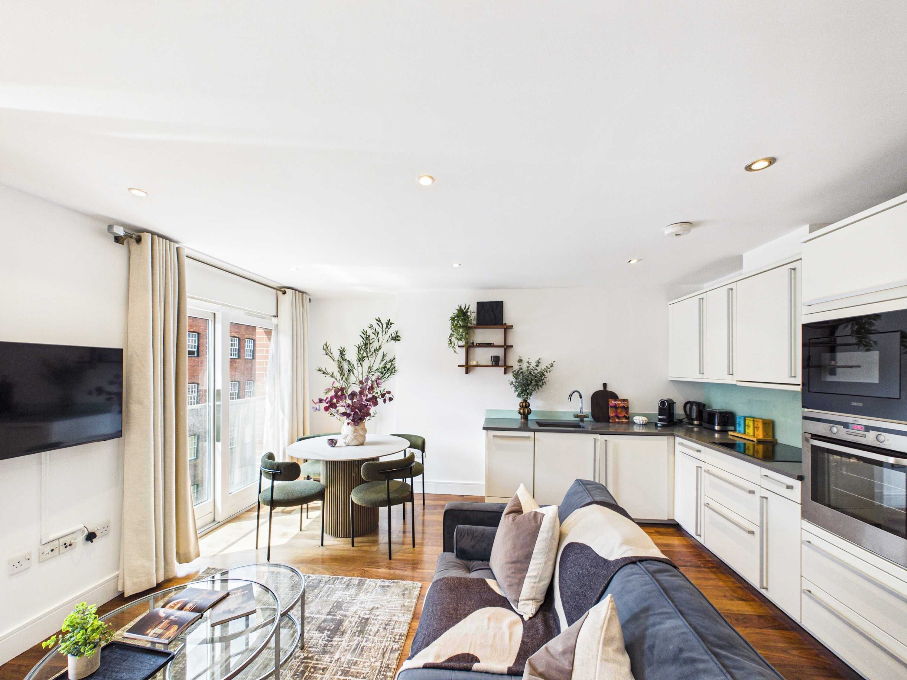A modern apartment with a living room, dining area, and kitchen. There is a sofa, a dining table with chairs, and a kitchen counter with appliances.