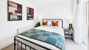 Ein modernes Schlafzimmer mit Bett, Nachttischlampen, einem Beistelltisch und abstrakter Wandkunst.
