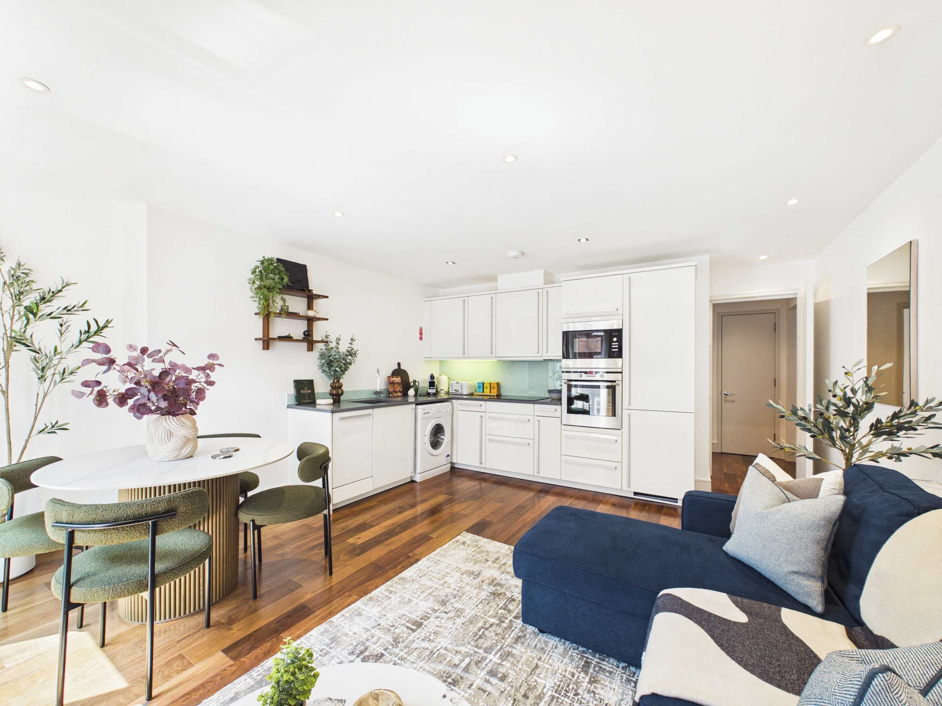 A modern kitchen with white cabinets, a built-in oven, and a microwave. There is a dining area with a round table and chairs, and a blue sofa in the living room.