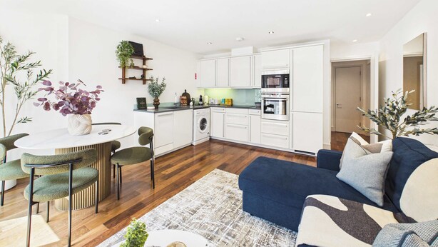 A modern kitchen with white cabinets, a built-in oven, and a microwave. There is a dining area with a round table and chairs, and a blue sofa in the living room.