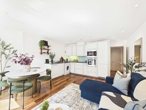 Eine moderne Küche mit weißen Schränken, einem eingebauten Ofen und einer Mikrowelle. Ein Essbereich mit rundem Tisch und Stühlen, und ein blaues Sofa im Wohnzimmer.