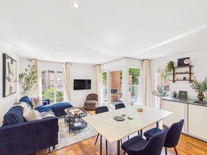 A modern living room with a sofa, a dining table, and chairs. There is a TV mounted on the wall, and a plant on the table. - Westminster Spacious Apartment with Balcony (Greater London)