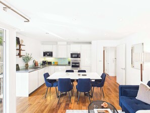 A modern kitchen with white cabinets, stainless steel appliances, and a dining area with a white table and blue chairs. - Westminster Spacious Apartment with Balcony (Greater London)