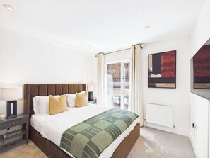 A hotel room with a bed, a nightstand, a lamp, a radiator, and a window with curtains. - Westminster Spacious Apartment with Balcony (Greater London)
