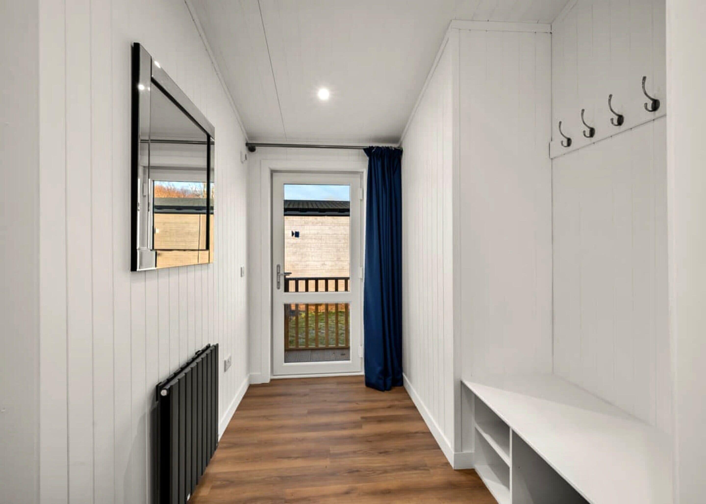 A hallway with a wooden floor, white walls, a radiator, a mirror, a door with a view to the outside, and a blue curtain.